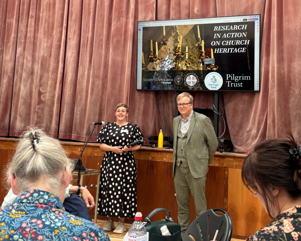 Lizzie (in a black and white polka dot dress) stands behind a microphone off centre of the frame, with DJ (in a three piece green tweed suit) stands next to her with his hands behind his back. Behind them is the stage which faded red velvet curtains, and a screen behind their heads with a slide saying "research in action on church heritage" and image of a chandelier. At the foreground of the picture are the back of the heads of audience members - two women are either side. On the left is a lady with grey hair in an up do, with blue patterned shirt and on the right is lady with brown hair and a dark shirt.