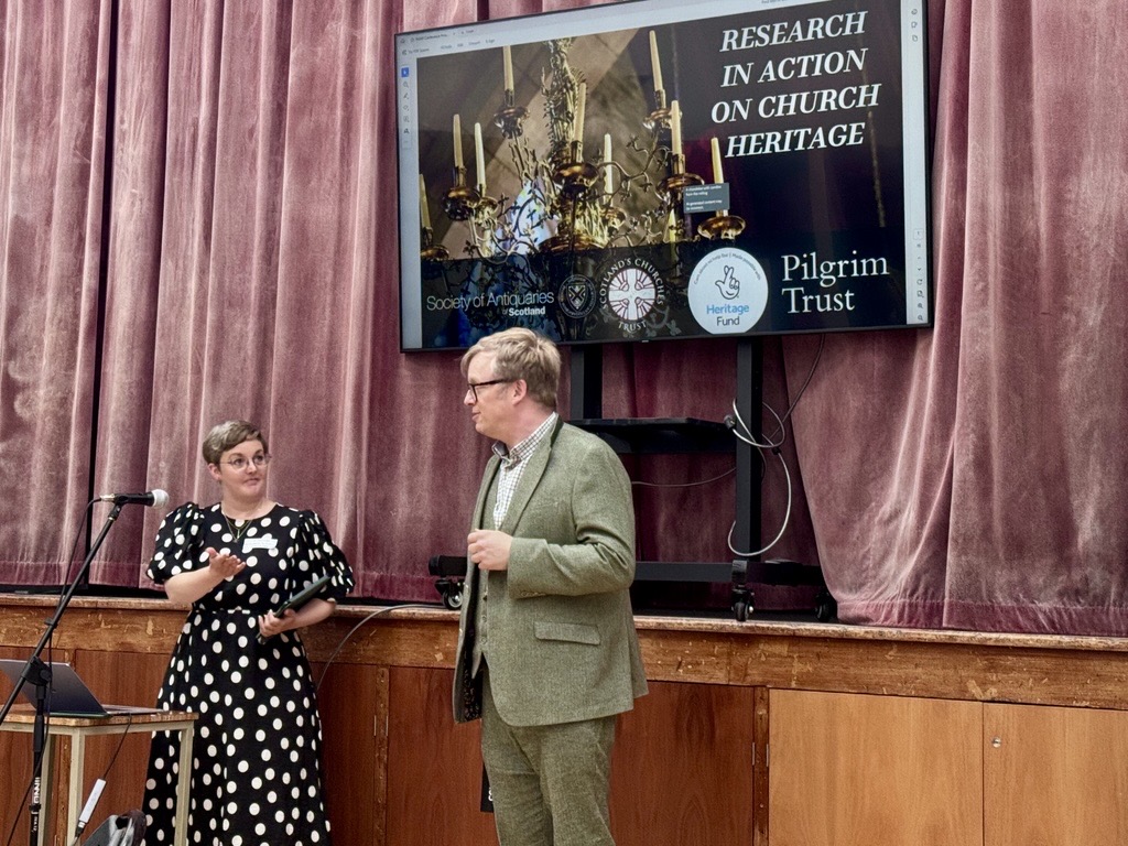 Lizzie and DJ in front of a stage and faded red velvet curtain with a screen behind them. Lizzie is in black and white polka dot dress and is gesturing to DJ on the right who wears a green tweed suit and glasses