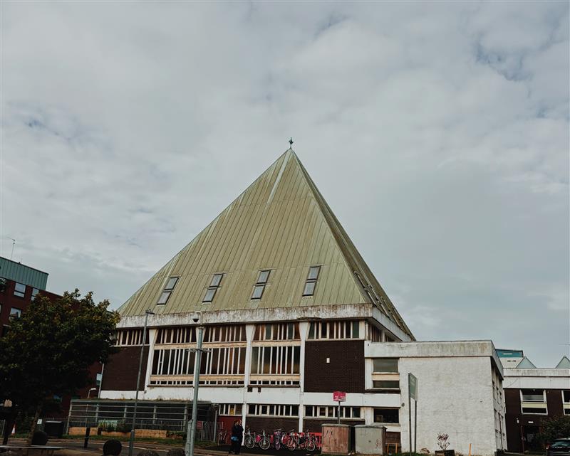 Photo of the Pyramid building, which is in the centre of the frame. The pointed roof of the building is green with four small windows at the bottom. The roof sits on a brown square building with white columns and panels at the windows. Next to this is a concrete square building. The sky takes up 2/3 of the frame and is mostly clouds with some blue. At the far left of the frame is a tree, coming to the height where the roof starts.