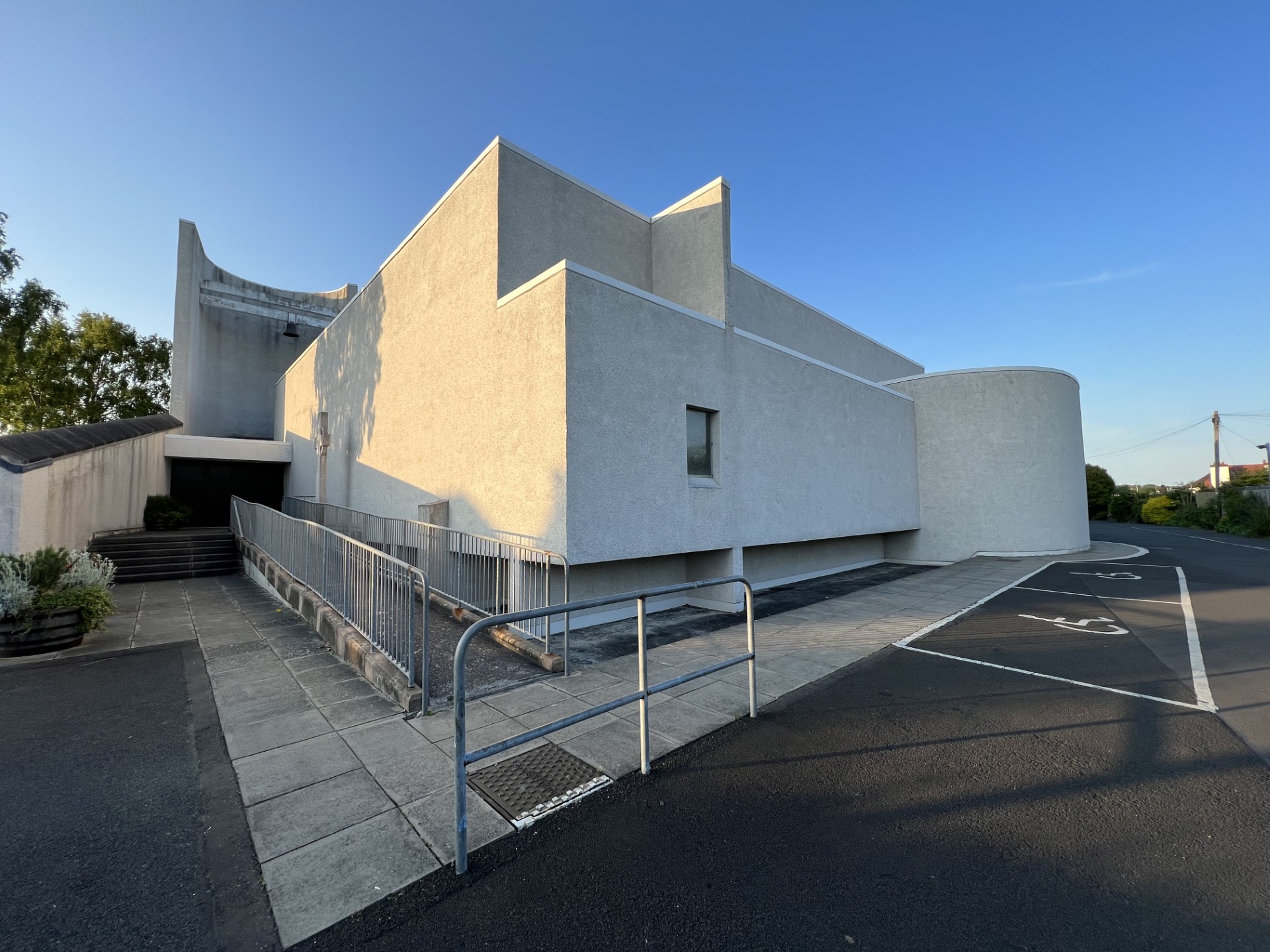Modern white concrete building with geometric shapes and a curved corner wall, viewed from a car park at sunset.