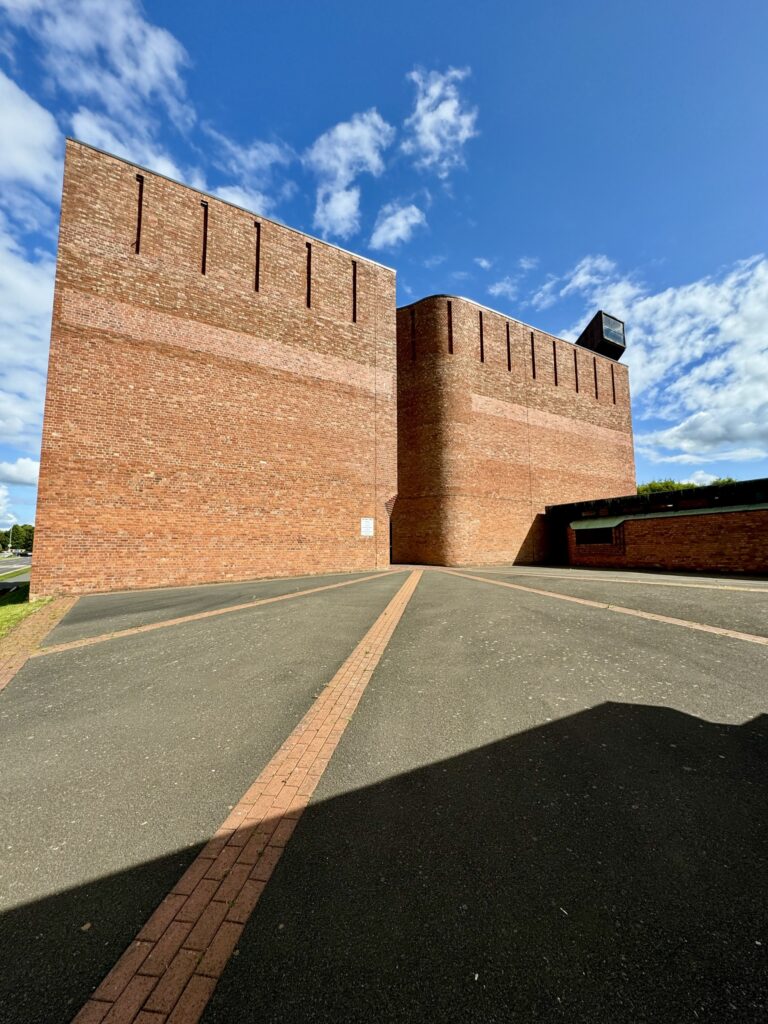 Close-up view of a red-brick building with tall rectangular walls and narrow vertical openings, photographed from a paved walkway.