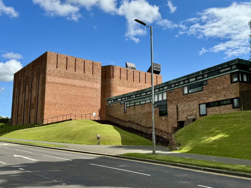 Large red-brick building with tall, windowless walls and a smaller connected structure on a grassy hill under a blue sky.