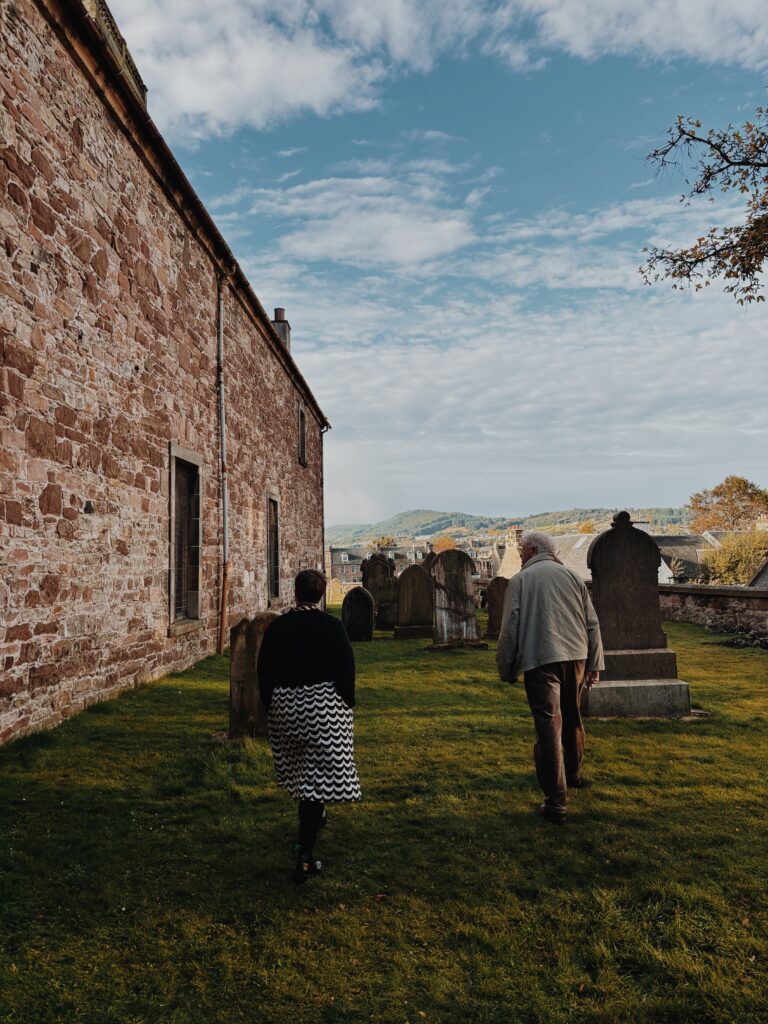 Two figures with their backs to camera walking through the church graveyard, with the church to the left

