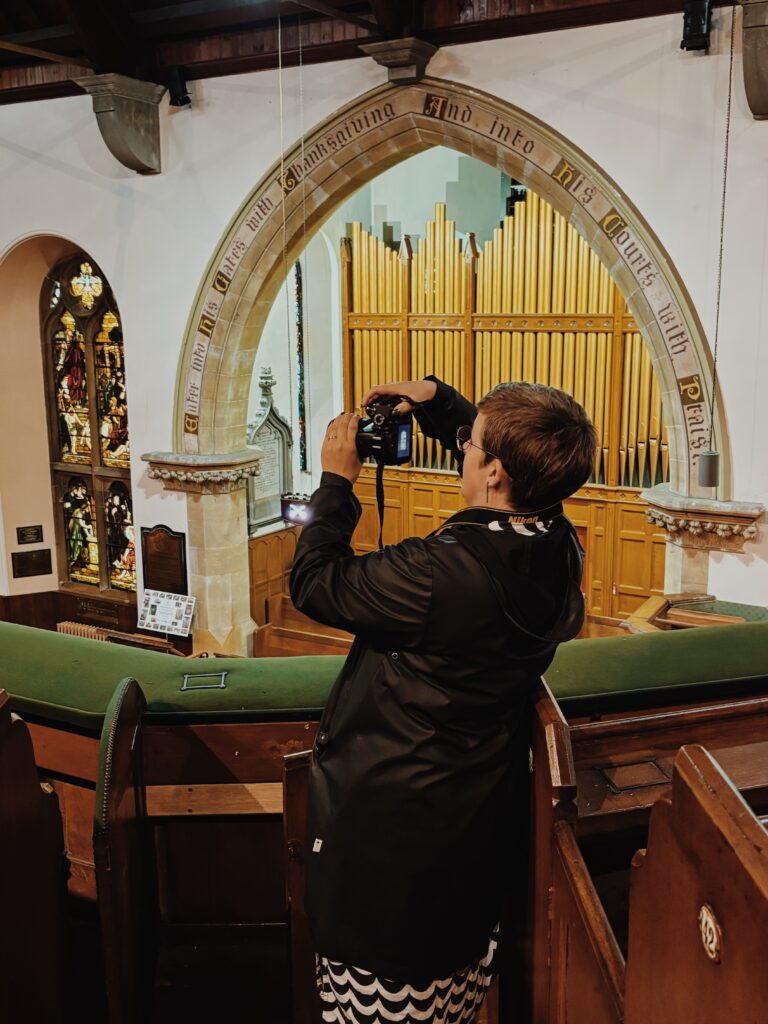 Lizzie standing side on in the middle of the frame, on the balcony of the interior of the church. She is taking a photo with camera pointing to the left