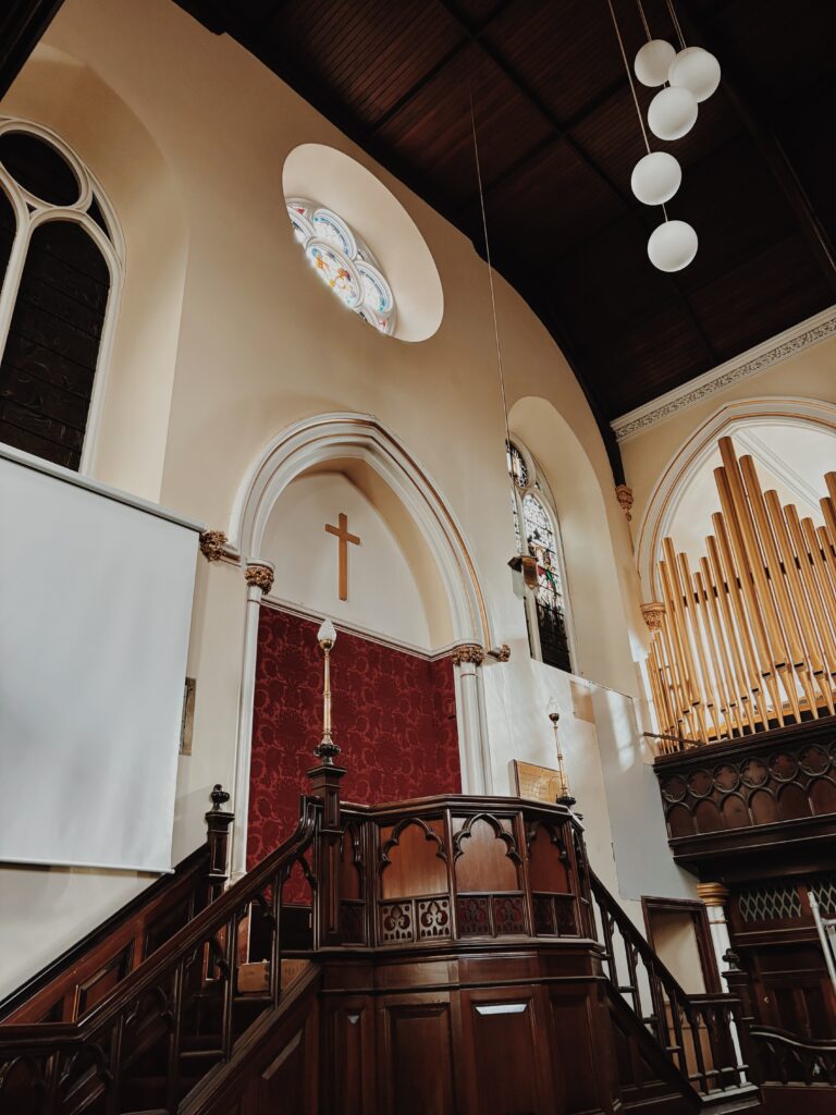 The wooden stairs to the pulpit of East Church, with the organ to its side, and above is a gold cross and a stained glass window