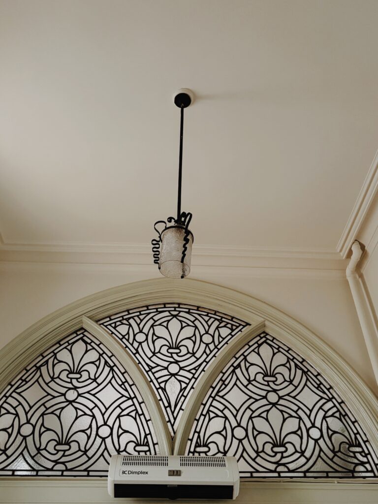 The black and white patterned glass of an arched window, with a light hanging in front which has a intricate iron detailing